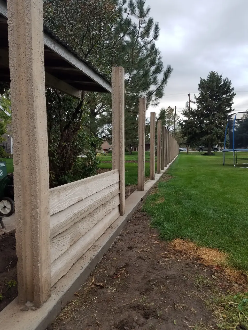 Precast concrete fence posts with wood-grain panels partly installed along a lawn